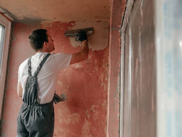 Young man builder dressed in work clothes overall applying putty mortar on the balcony wall with a spatula.