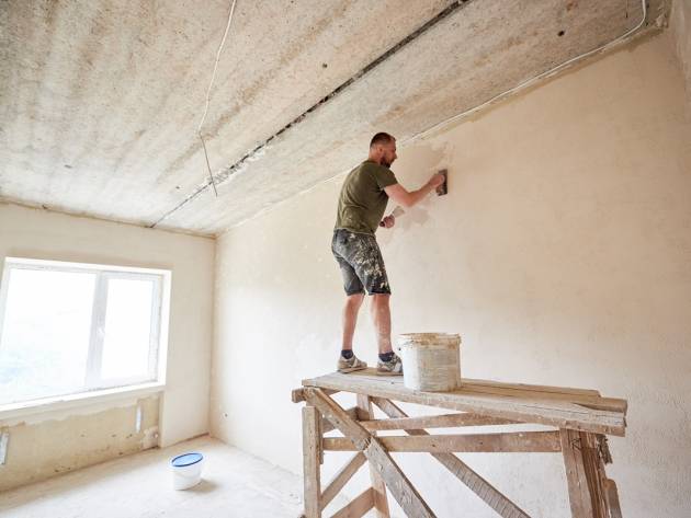 Young man is standing on wooden stand and working with spatula with plaster at the wall. Builder man is standing on a wooden stand and working with a spatula with plaster on the wall against a window. Puttying the walls indoors. A guy with a beard in a t-shirt and jeans is smeared paint